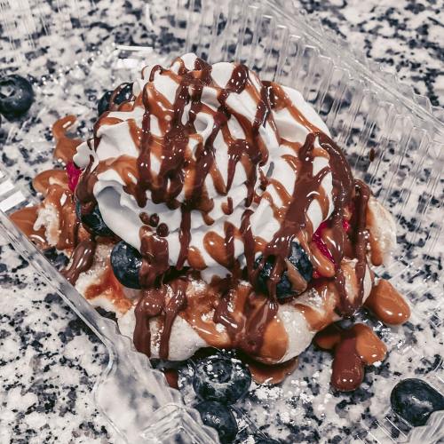 A dessert topped with whipped cream, chocolate drizzle, blueberries, and strawberries in a clear container.