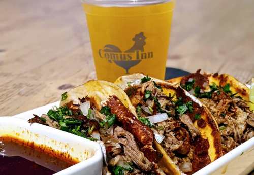Three tacos filled with beef, onions, and cilantro, served with a side of sauce and a drink in a branded cup.
