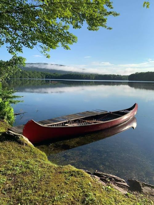 A red canoe rests on the shore of a calm lake, surrounded by lush greenery and misty mountains in the background.