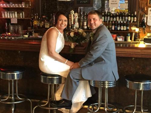 A bride and groom sit on bar stools, smiling at each other in a cozy bar setting, surrounded by bottles and decor.