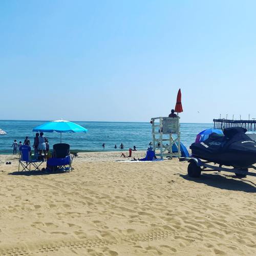 A sunny beach scene with umbrellas, chairs, and people enjoying the water near a lifeguard station and pier.