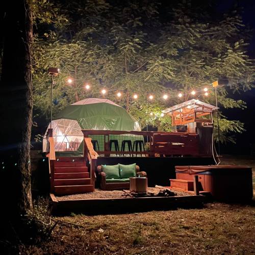 A cozy campsite at night with a green tent, wooden deck, string lights, and a fire pit surrounded by trees.