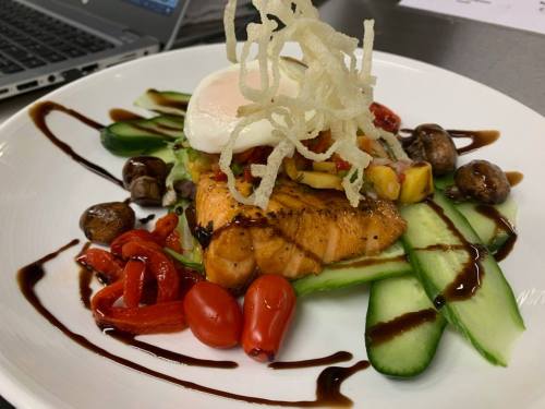 A beautifully plated dish featuring salmon, a poached egg, vegetables, and a drizzle of sauce, garnished with crispy strands.