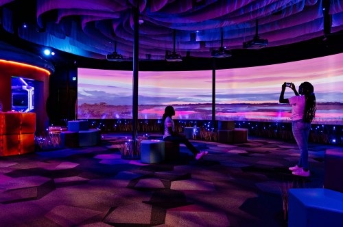 Two people in a colorful, modern room with large screens displaying a scenic landscape. Soft lighting and seating are visible.