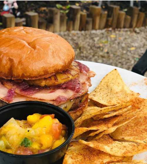 A stacked sandwich with ham and pineapple on a bun, served with tortilla chips and a small bowl of salsa.