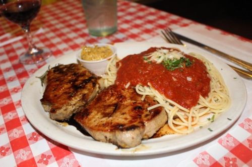 A plate of grilled meat with spaghetti topped in marinara sauce and a side of mustard, set on a checkered tablecloth.