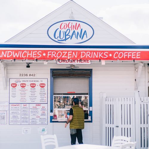 A person approaches a food stand with a sign for sandwiches, frozen drinks, and coffee, labeled "Cocina Cubana."