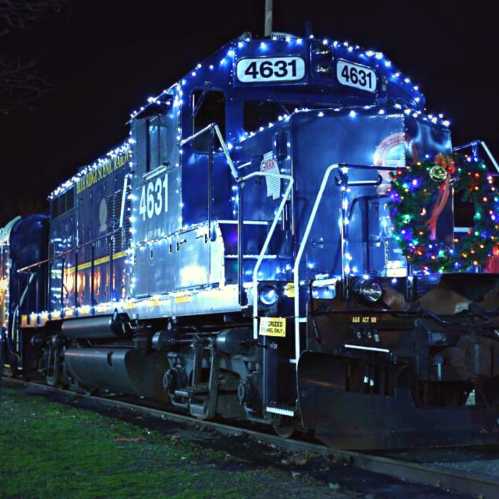 A brightly lit blue train decorated with festive lights and a wreath, parked at night.