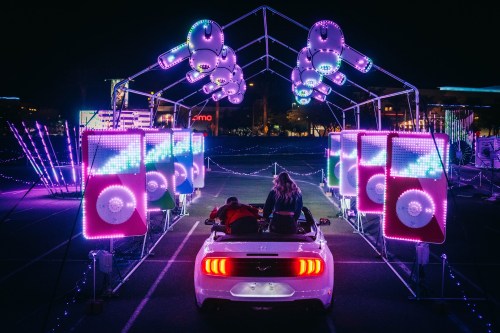 A couple sits in a white convertible under colorful, illuminated arches and speakers at a nighttime event.