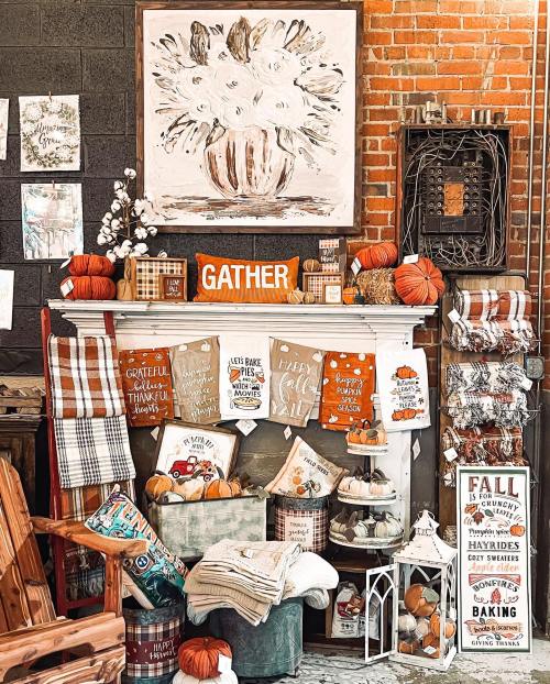 Cozy fall-themed display with pumpkins, blankets, and a "GATHER" sign on a rustic mantel against a brick wall.