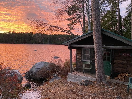 A cozy cabin by a lake at sunset, with vibrant orange and pink skies reflecting on the water.