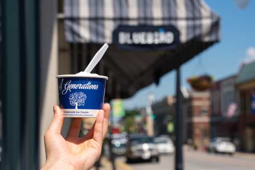 A hand holds a blue ice cream cup in front of a shop with a striped awning on a sunny day.