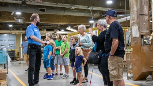 A group of children and adults listen to a guide in a factory setting, with machinery in the background.
