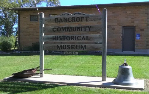 Sign for Bancroft Community Historical Museum, established 2003, with a bell and wheelbarrow nearby on a grassy area.