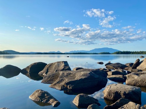 A serene lake scene with smooth rocks in the foreground, reflecting clouds and mountains under a clear blue sky.