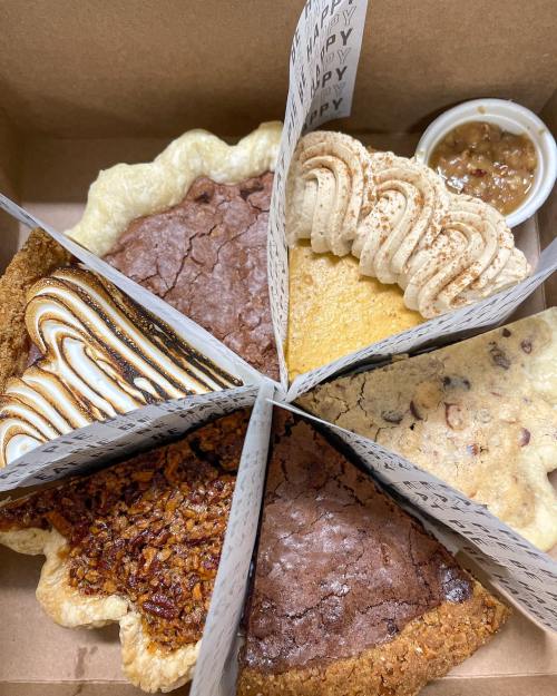A box containing six assorted pies, including chocolate, pecan, and pumpkin, with a small container of sauce.
