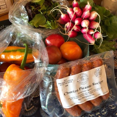 A variety of fresh vegetables including peppers, tomatoes, radishes, and chorizo sausage on a patterned tablecloth.