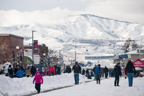 A snowy street scene at Lake Chelan Winterfest, with people walking and mountains in the background.