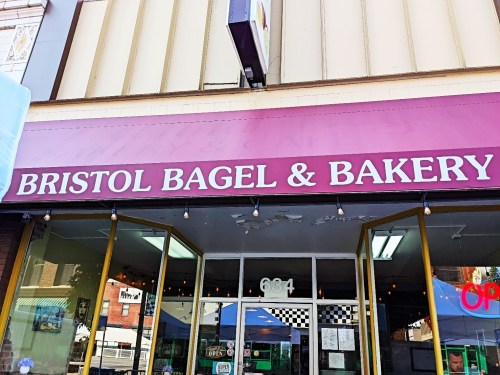 Sign for Bristol Bagel & Bakery, featuring a purple awning and large white lettering. Storefront visible with open sign.
