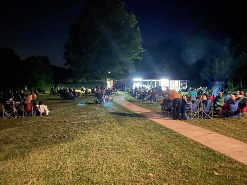 A crowd of people sitting in chairs outdoors at night, facing a lit area with a building and trees in the background.