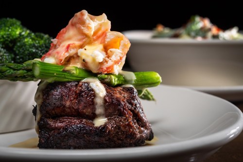 A gourmet dish featuring a grilled steak topped with lobster, asparagus, and a creamy sauce, served with broccoli.