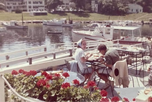 A couple enjoys a meal at an outdoor café by the water, surrounded by boats and blooming flowers.