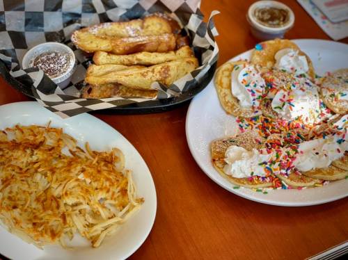 A plate of crispy hash browns, a basket of French toast sticks, and a plate of colorful pancakes with whipped cream and sprinkles.