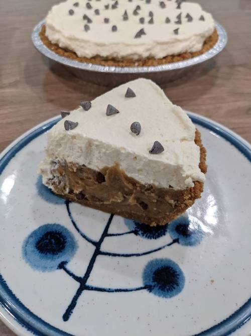 A slice of pie with a creamy topping and chocolate chips, served on a decorative plate, with a whole pie in the background.