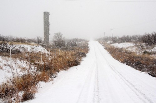 A snowy dirt road stretches into the distance, flanked by sparse vegetation and a tall structure in the background.