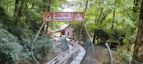 Sign reading "Slow! Track ends in 100ft" above a winding roller coaster track surrounded by trees and a rustic building.