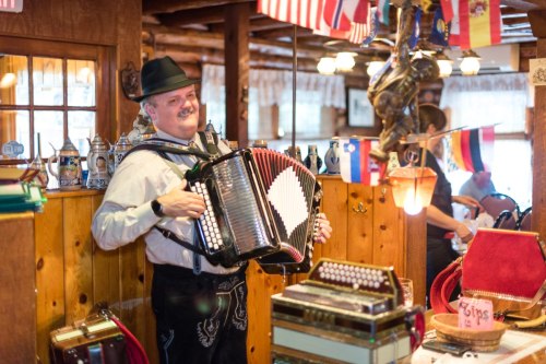 A man in traditional attire plays the accordion in a cozy, decorated restaurant with flags and wooden decor.