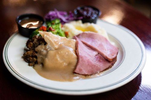 A plate of food featuring sliced ham, mashed potatoes, stuffing, and mixed vegetables with gravy and a side of sauce.