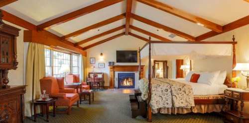 Cozy hotel room with a four-poster bed, seating area, fireplace, and wooden beams on the ceiling. Warm and inviting atmosphere.
