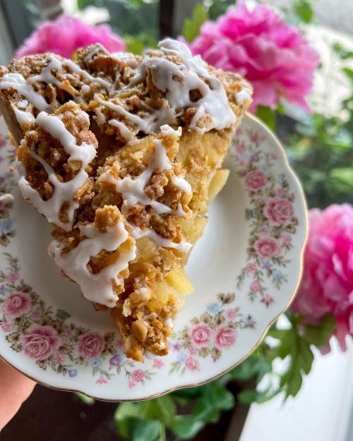 A slice of apple crumble pie drizzled with icing, served on a floral plate, with pink peonies in the background.