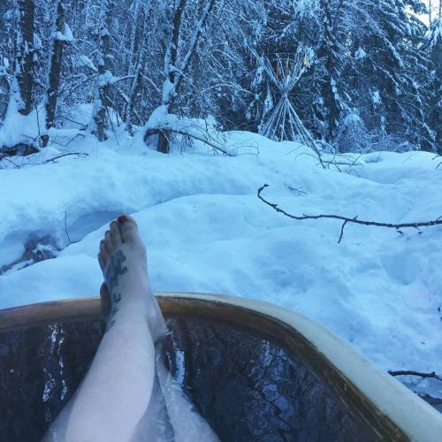 A foot with a tattoo rests on the edge of a wooden tub, surrounded by snow-covered trees and a winter landscape.