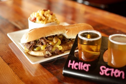 A hearty sandwich filled with beef and peppers, served with a side of macaroni salad and two small glasses of beer.