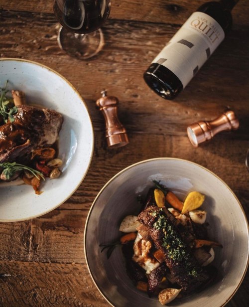 A top-down view of two gourmet dishes, a glass of red wine, and wooden pepper mills on a rustic table.