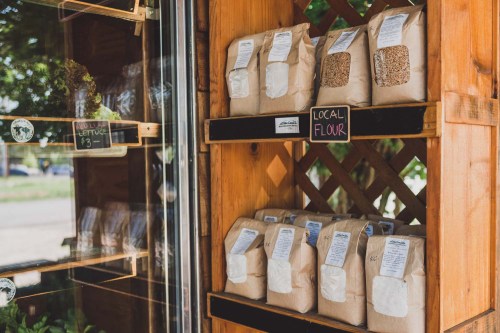 Bags of local flour and grains displayed on wooden shelves outside a market, with a sign reading "Local Flour."