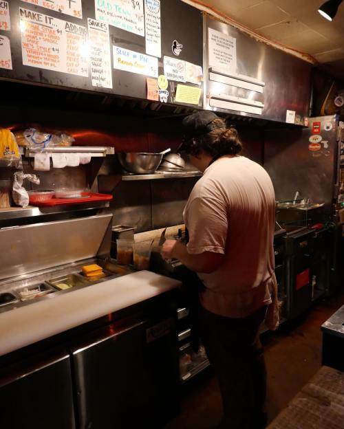 A person prepares food in a busy kitchen, surrounded by stainless steel equipment and handwritten menus.