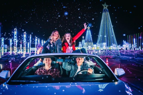 Four friends joyfully pose in a car, surrounded by colorful holiday lights and decorations at night.