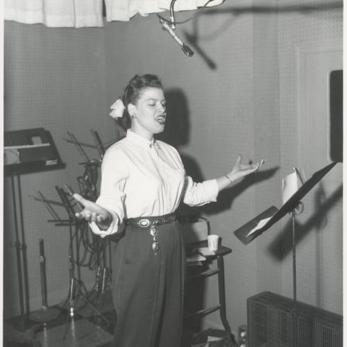 A woman sings passionately in a recording studio, with a microphone overhead and music stands in the background.