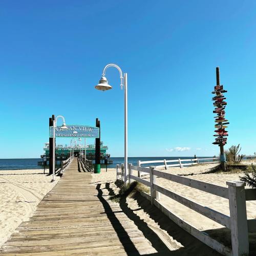 A sunny beach scene featuring a fishing pier, sandy path, and decorative pole against a clear blue sky.