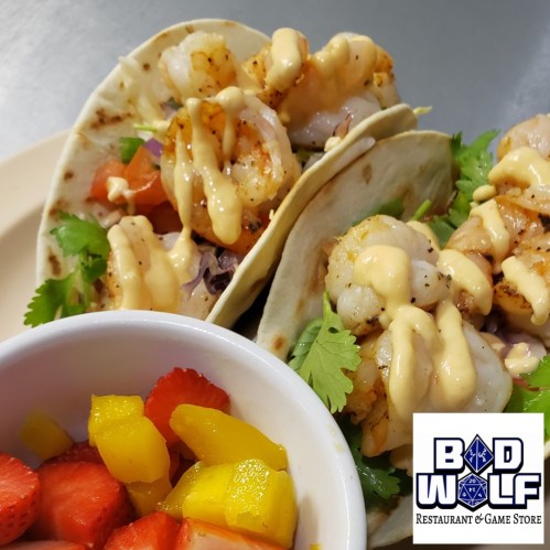 Three shrimp tacos topped with lettuce and sauce, served with a side of fresh fruit salad. Logo of Bad Wolf Restaurant.