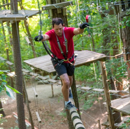 A man in a red shirt navigates a rope course high in the trees, using safety harnesses and equipment.