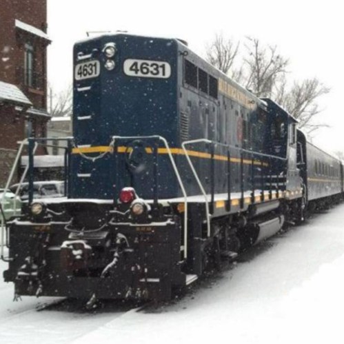 A blue and yellow train engine parked on a snowy track, surrounded by trees and buildings in winter.