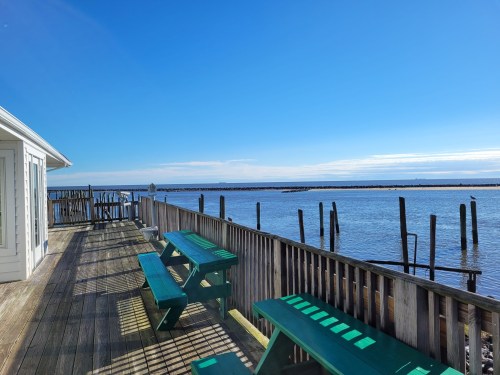 A wooden deck with green picnic tables overlooks a calm waterway under a clear blue sky.