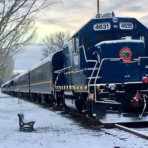 A blue train engine and passenger cars on snowy tracks, with trees and a bench in the background.