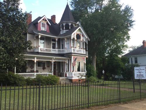 A large, historic Victorian-style house with a turret, surrounded by a green lawn and a black fence.