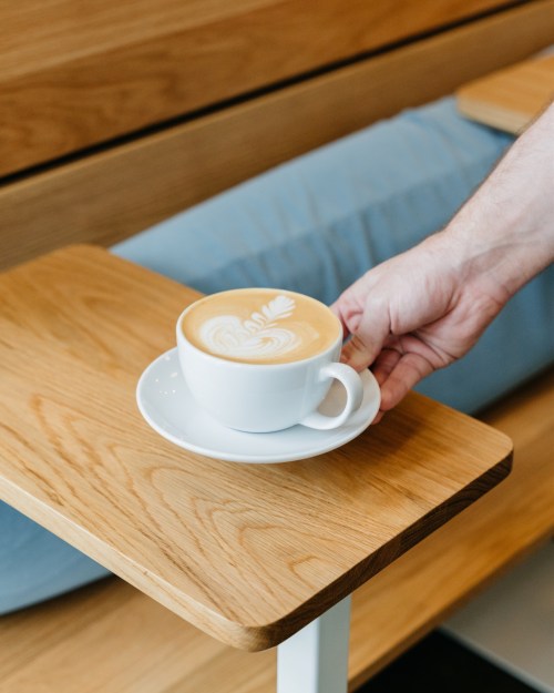 A hand reaching for a latte with latte art on a white cup, resting on a wooden table.