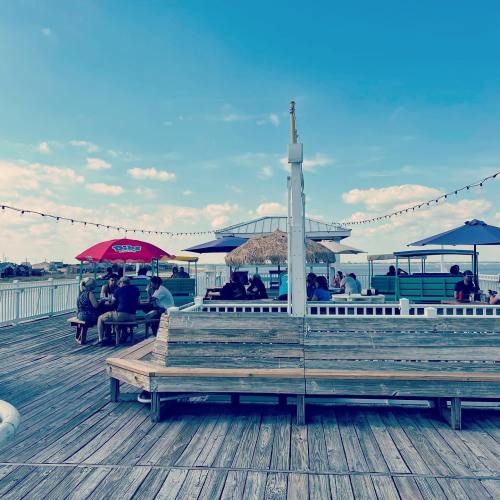 A wooden pier with tables and umbrellas, people dining, and a clear sky in the background.